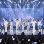 콘서트 무대 위에서 조명을 받으며 객석을 향해 서 있는 아이돌 그룹의 모습